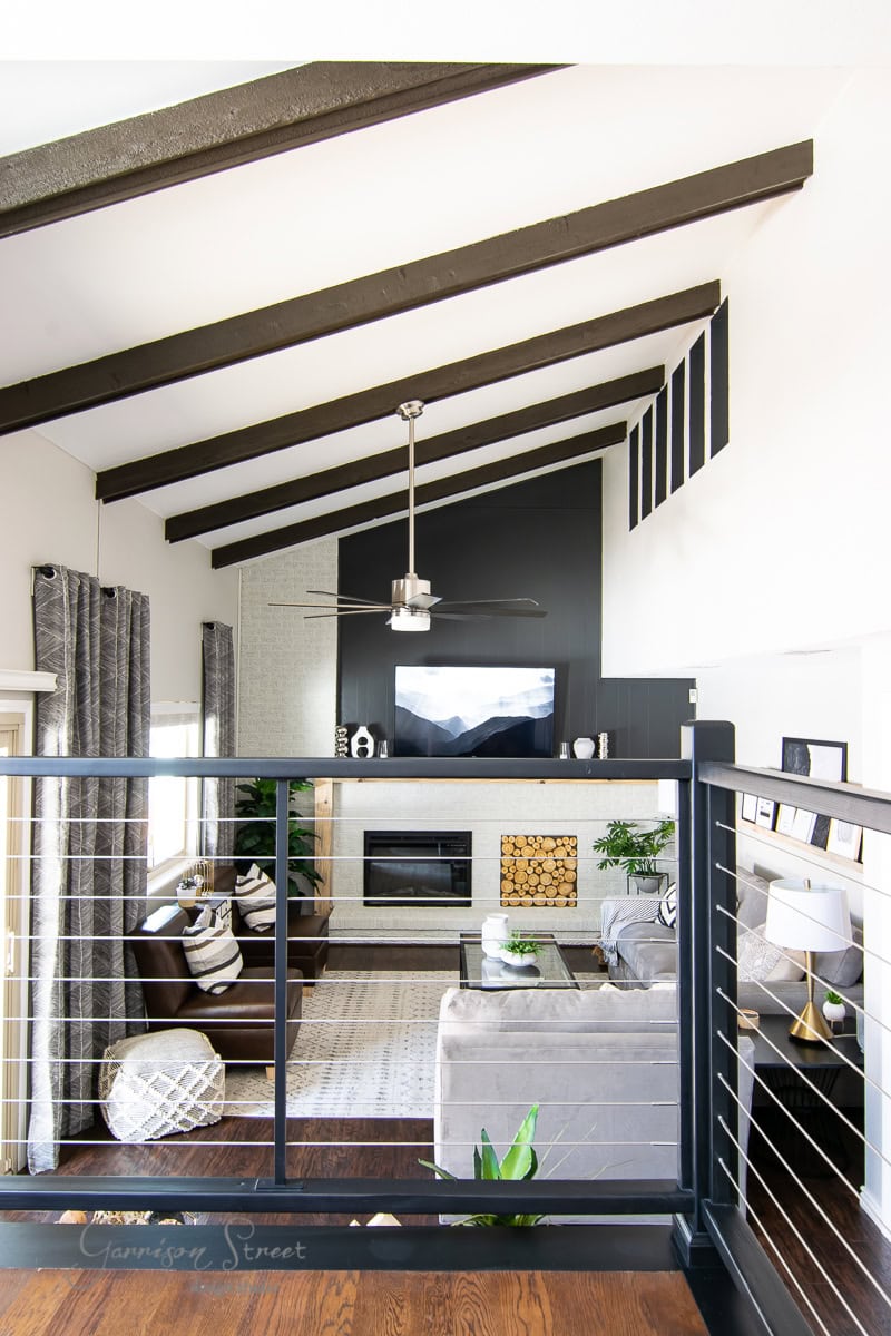 A modern living room with a high ceiling and exposed beams, embodying the best of comfort in 2021. Features include a gray sectional sofa, a fireplace with a TV above, a coffee table, and decorative accents. Natural light filters through large windows, highlighting an inviting space.