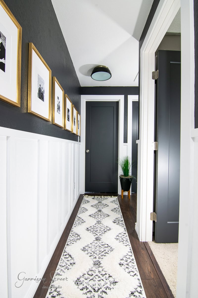 A modern hallway, reminiscent of 2021's best designs, features white wainscoting with black upper walls adorned with framed photos. A sleek black door stands at the end, accented by a geometric-patterned runner. A potted plant near the door adds a refreshing touch of greenery.
