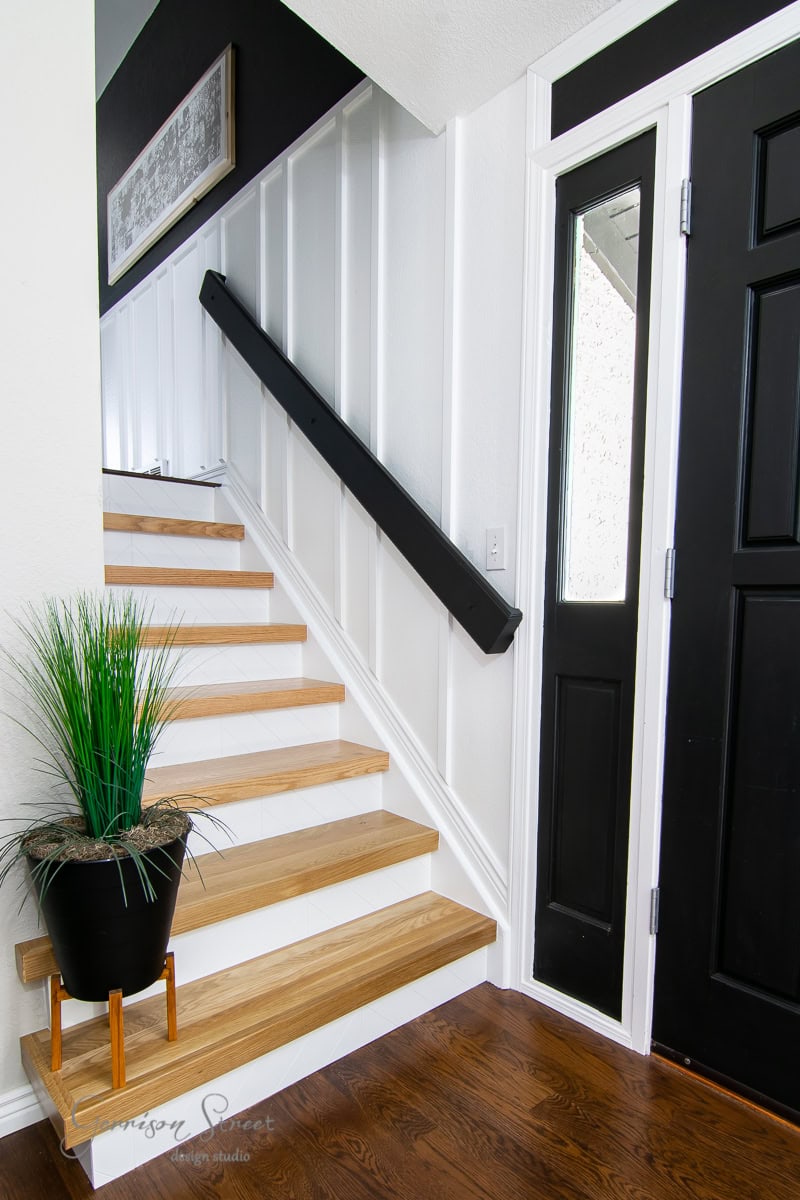 Voted "Best of 2021," this modern staircase boasts light wood steps against a pristine white wall, complemented by a sleek black handrail. Adjacent is a striking black front door with a vertical window. A potted plant on a step adds vitality to the elegant space.