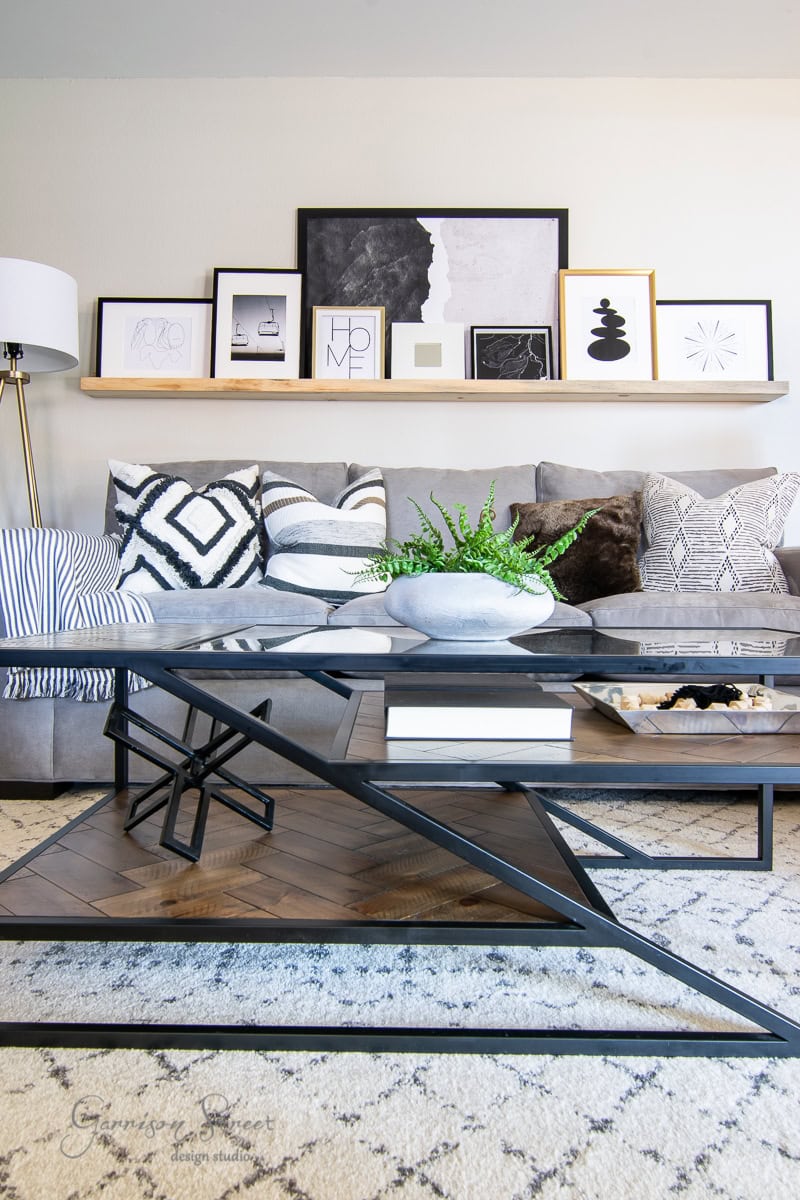 A modern living room features a gray sofa adorned with patterned cushions. A sleek, angular coffee table, worthy of a 2021 Best of design list, holds books, a tray, and a plant. Above, a shelf showcases framed artwork in various sizes and styles. A floor lamp stands to the side.