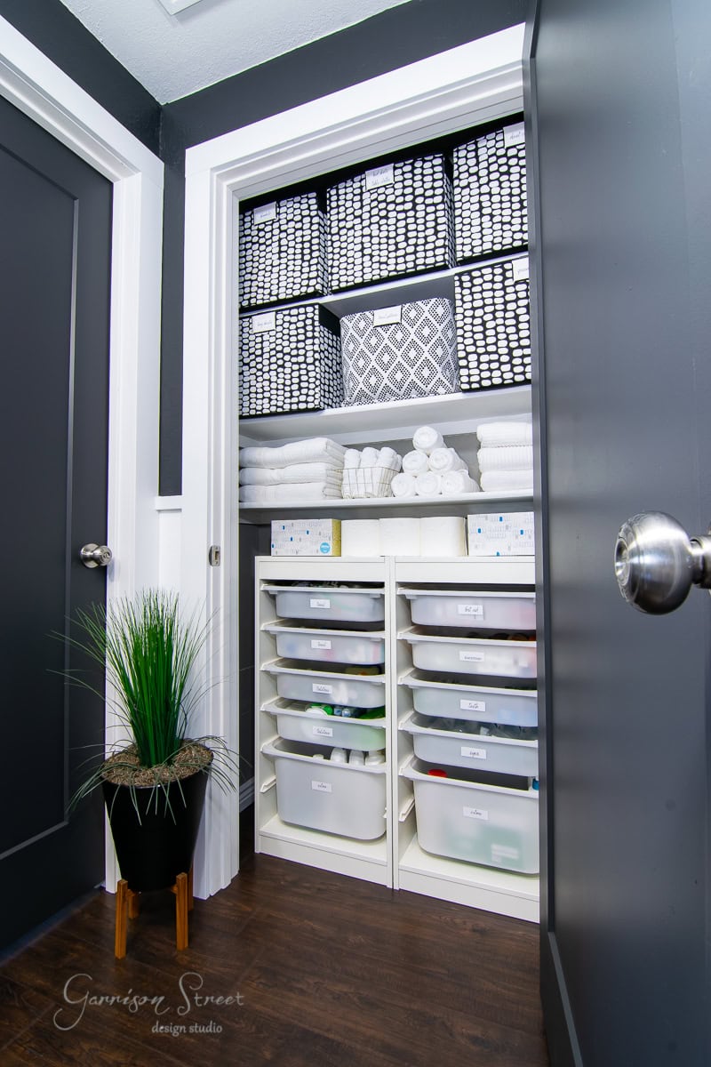 A neatly organized closet, named Best of 2021, features black and white baskets on top shelves, white folded towels in the middle, and transparent storage bins with labels on the bottom. A potted plant sits next to the open door, revealing a dark interior.