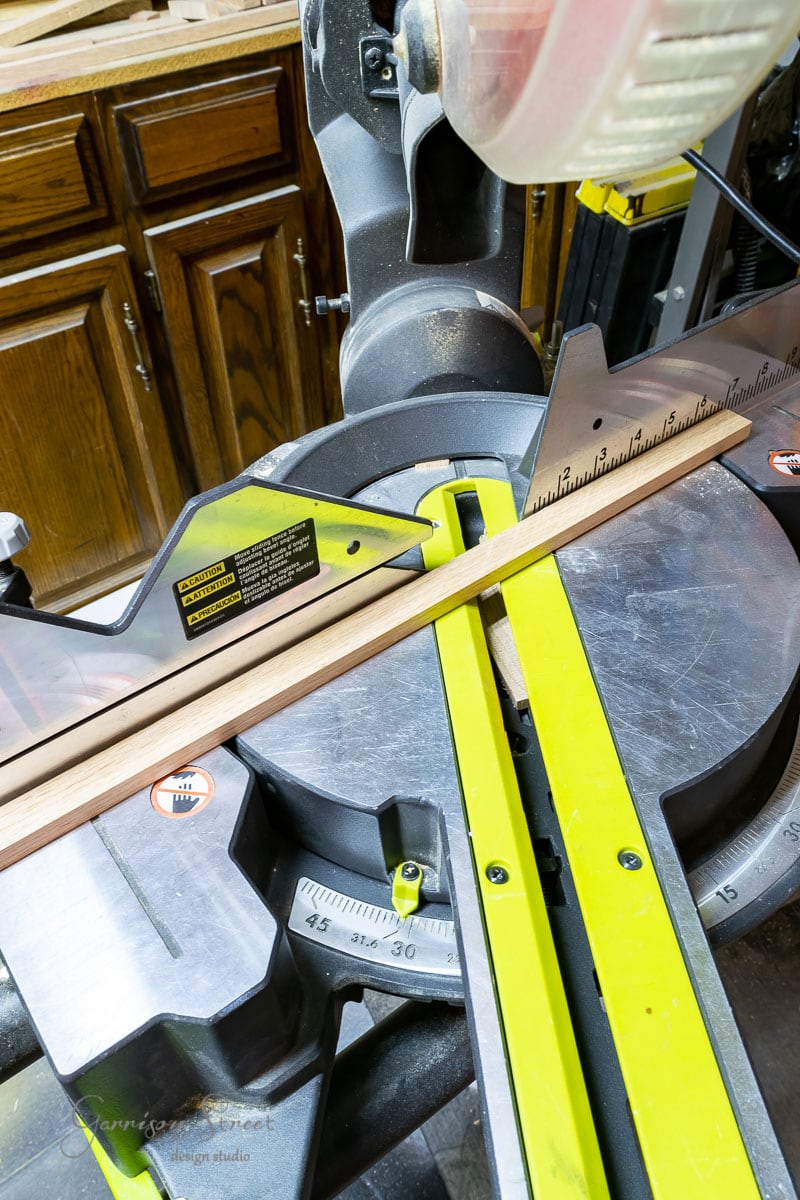 A miter saw is positioned on a workbench with a piece of wood ready to be cut, perhaps for crafting scrap wood Christmas trees. The circular blade is prominently visible, and measurement markings adorn the saw's base. Wooden cabinets are in the background.