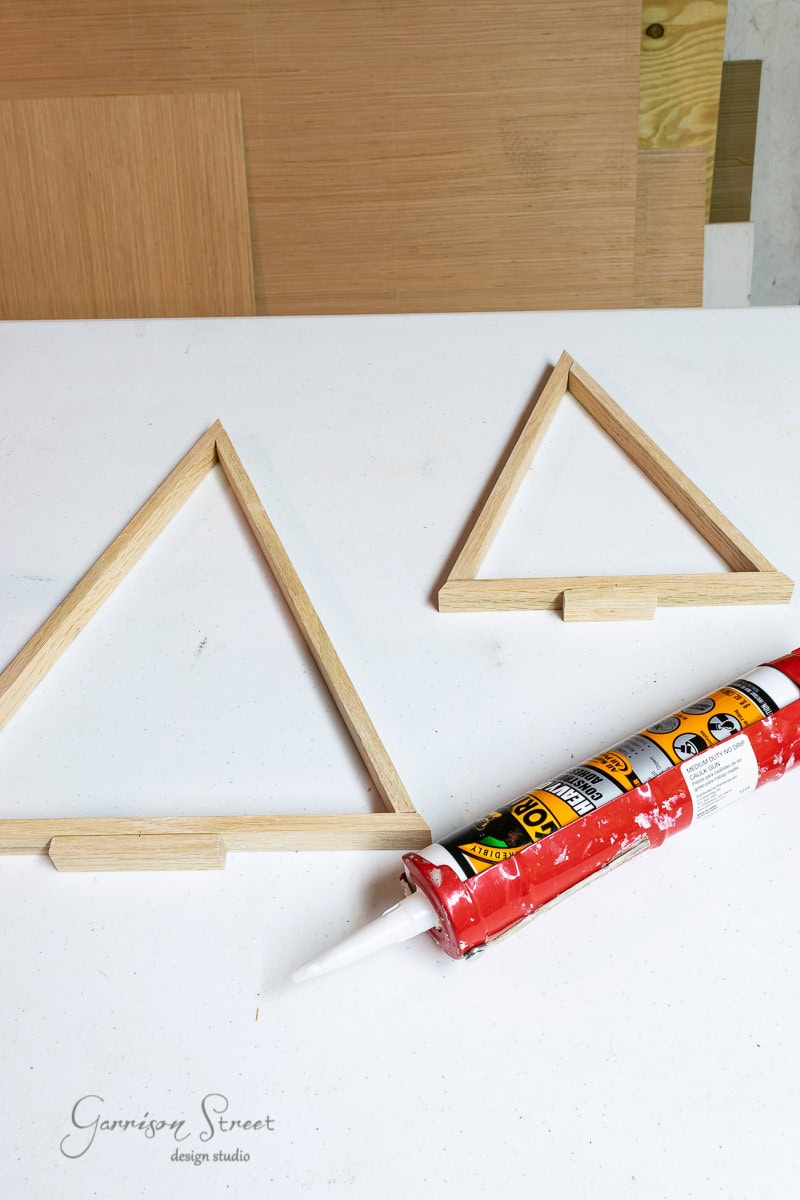 Two wooden triangular frames on a table beside a tube of adhesive, resembling the early stages of crafting scrap wood Christmas trees. The unassembled pieces lay neatly while light wood panels form the backdrop, setting the scene for a cozy DIY project.
