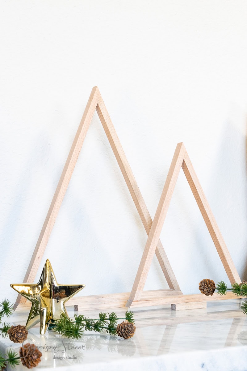 Scrap wood Christmas trees in triangular shapes are displayed on a marble surface. In front, a gold star ornament is surrounded by small pine cones and sprigs of greenery. The background is a plain white wall, enhancing the minimalist charm.