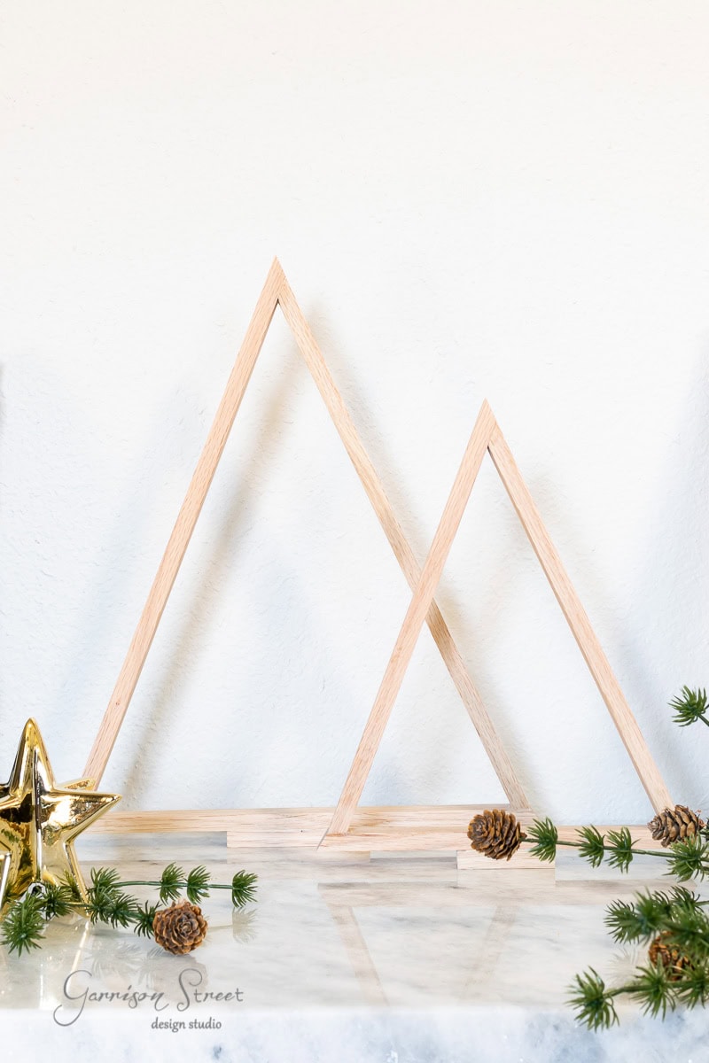 Wooden triangular sculptures, reminiscent of scrap wood Christmas trees, are arranged on a marble surface. Adorned with small pinecones and sprigs of green foliage, they create a festive display. A gold star ornament rests to the left against a plain white wall backdrop.