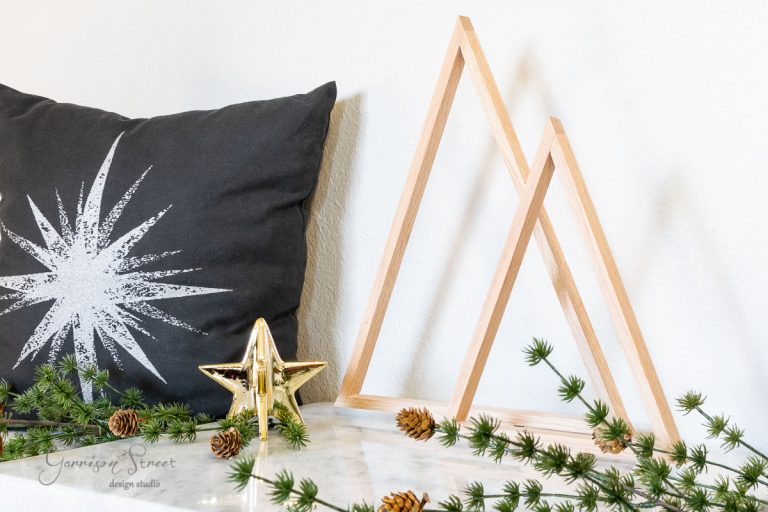 A decorative setup with a black pillow featuring a white starburst design, scrap wood Christmas trees, wooden triangular sculptures, a small golden star, and pine branches with cones on a marble surface.