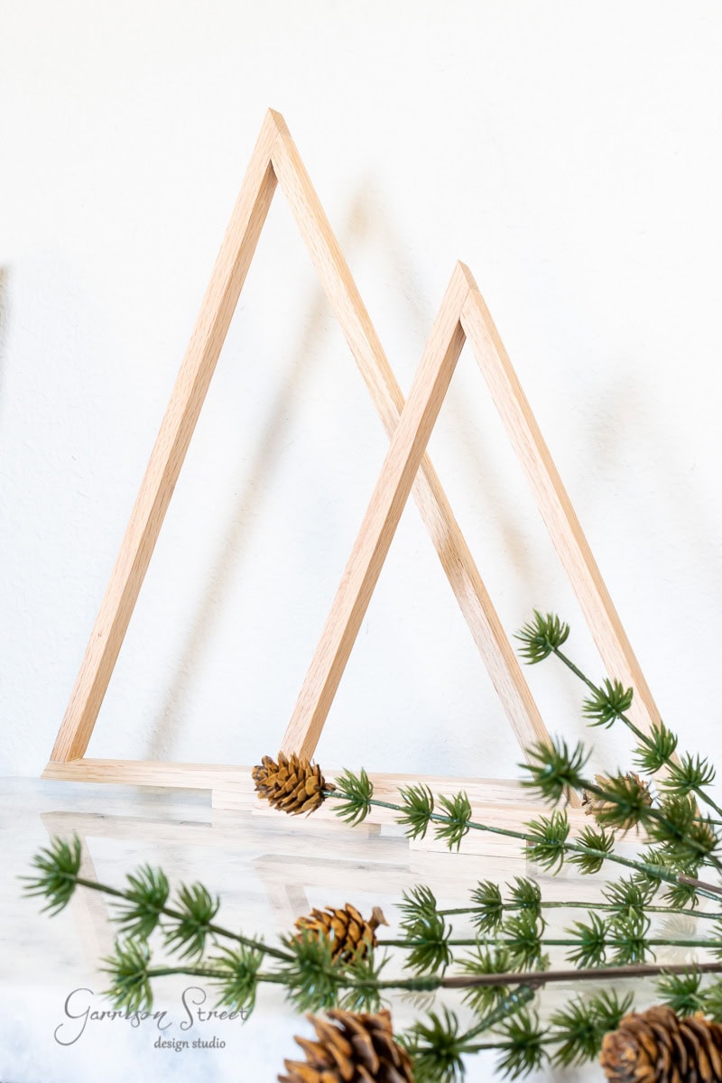 Wooden triangular frames, reminiscent of scrap wood Christmas trees, are arranged against a white background. Pine branches and small pinecones are artfully placed in front. The text "Garrison Street Design Studio" is visible in the bottom left corner.