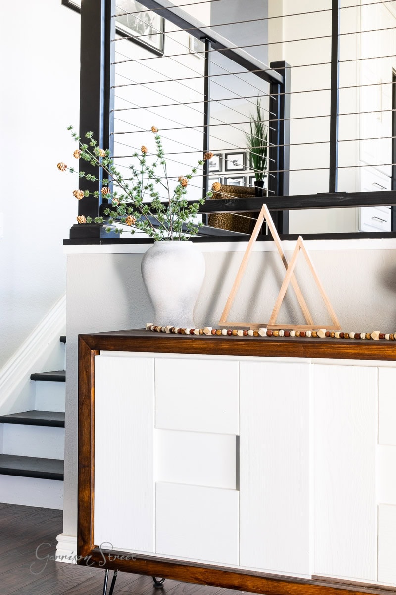Modern interior with a decorative white console table featuring a wooden frame. On top, there's a white vase with branches and a wooden triangular sculpture. A staircase with black railing is in the background, complemented by charming scrap wood Christmas trees, adding festive cheer.