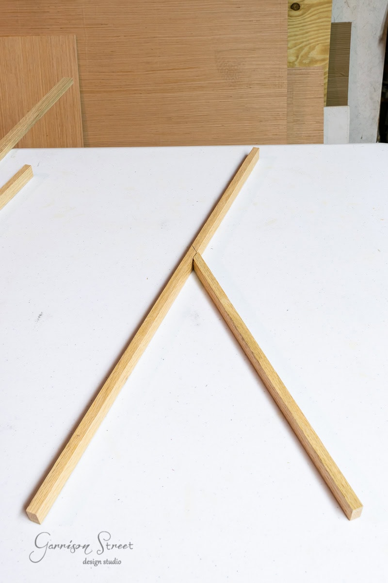 Two wooden strips are arranged on a white surface in a "V" shape, resembling scrap wood Christmas trees. In the background, several boards of various sizes lean against each other. A logo reading "Garrison Street Design Studio" is visible on the left.