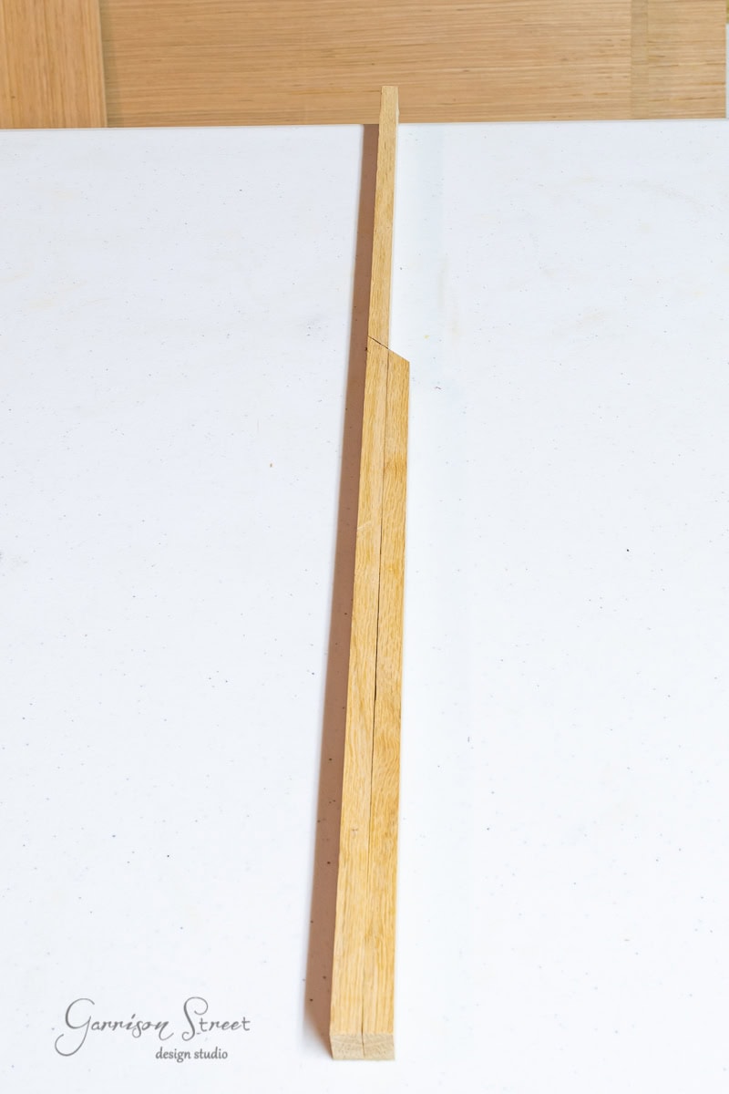 A long, thin piece of wood with a bevel cut rests on a white surface, accompanied by the faint logo "Garrison Street design studio" in the bottom left corner. Its simplicity evokes the charm of scrap wood Christmas trees.