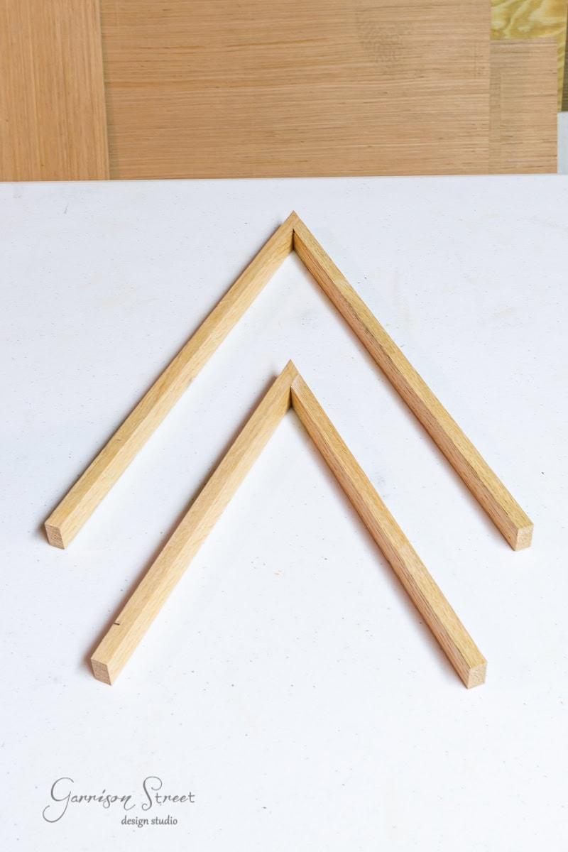 Two overlapping wooden angle frames form a chevron pattern on a white surface, reminiscent of scrap wood Christmas trees. Various shades create a background with subtle geometric shapes. A logo reads "Garrison Street design studio" in the bottom left corner.