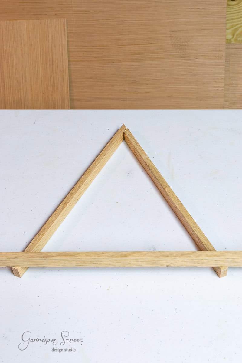 A wooden triangular frame crafted from scrap wood with a base support lies on a white surface. The background features various shades of wooden panels, reminiscent of rustic Christmas trees. The words "Garrison Street design studio" are visible in the bottom left corner.