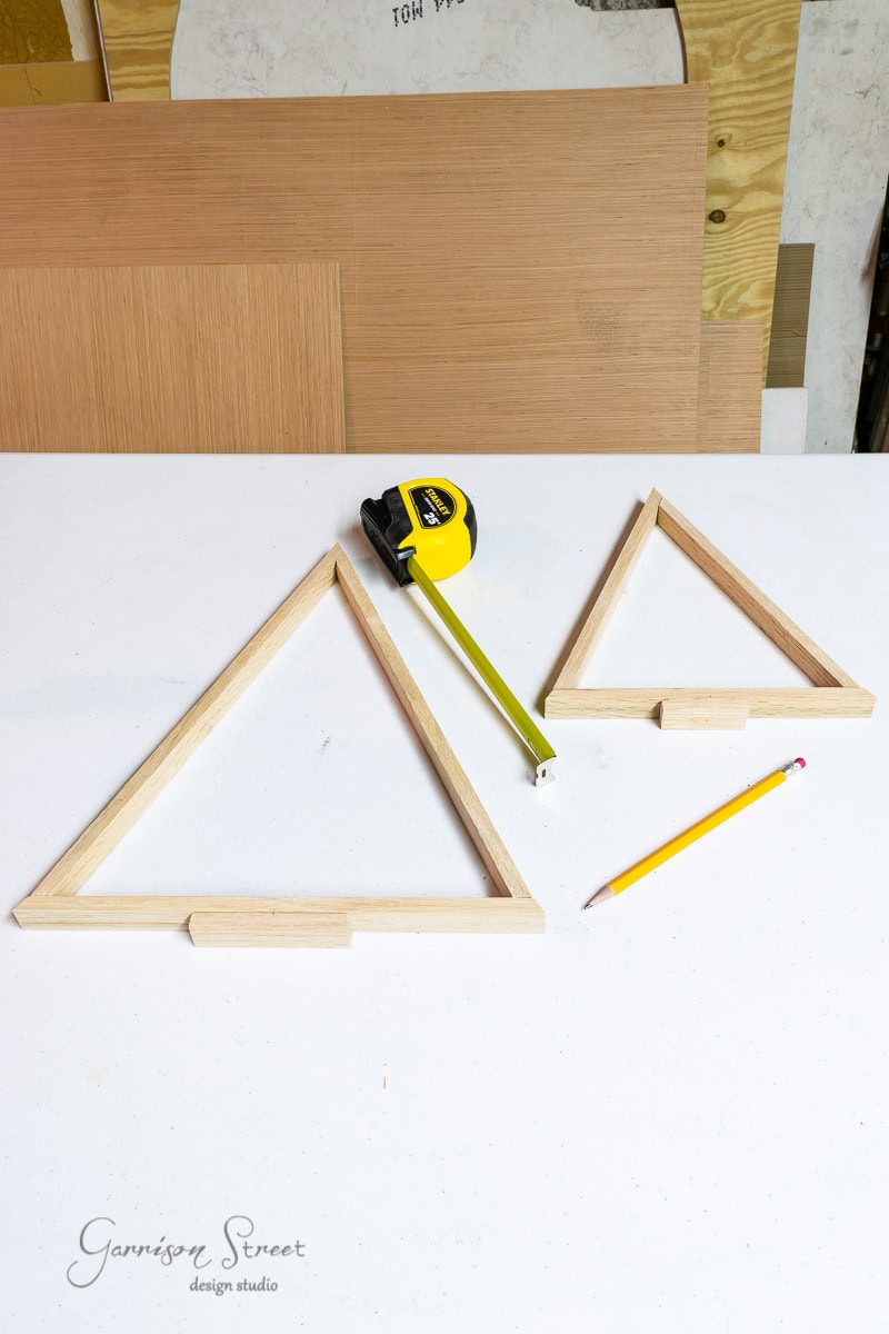 Two wooden triangular frames, a yellow measuring tape, and a pencil are neatly arranged on a white surface. In the background sits a stack of wood, hinting at the raw materials for crafting charming scrap wood Christmas trees.