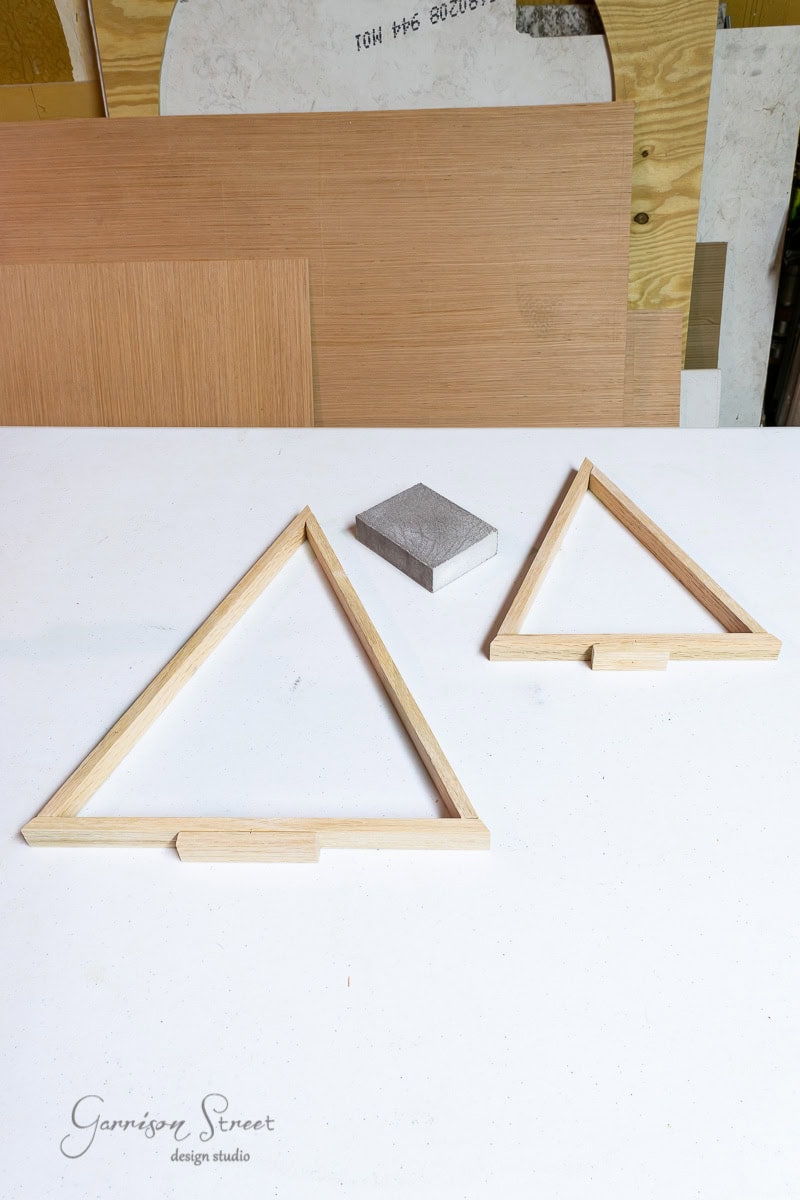 Two wooden triangles resembling scrap wood Christmas trees and a rectangular block rest on a white table. Wood panels are stacked in the background, suggesting a workspace, possibly related to crafting or carpentry.