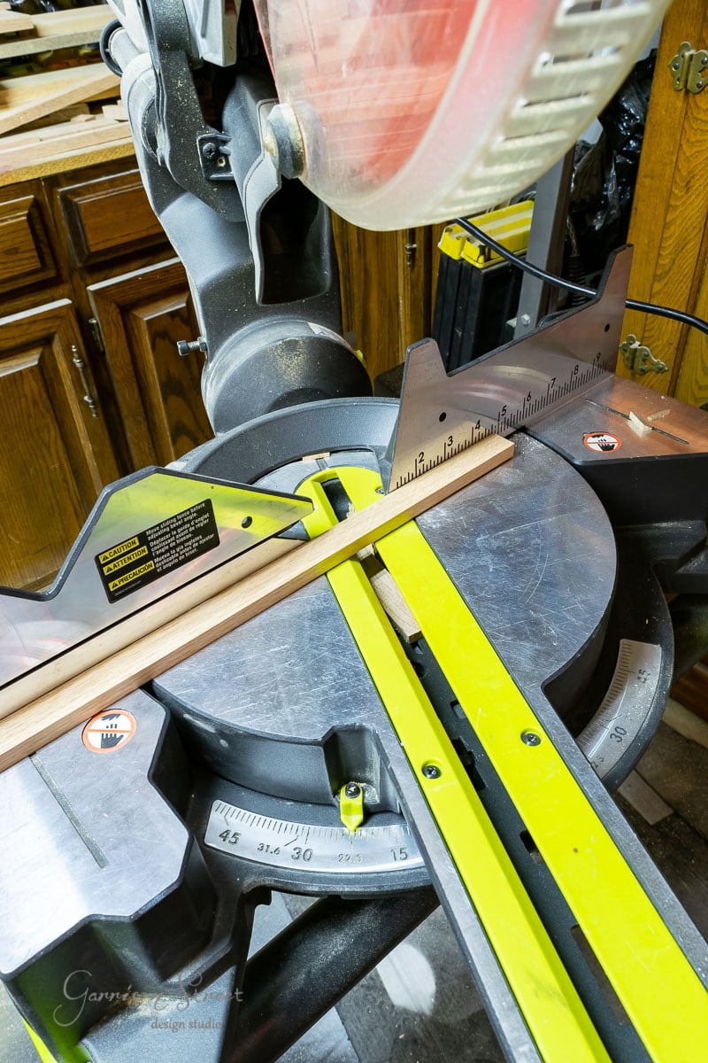A miter saw with a wooden board ready for trimming sits on the workbench, ideal for crafting scrap wood Christmas trees. Cabinets stand in the background while the saw's measurement guide is clearly visible.