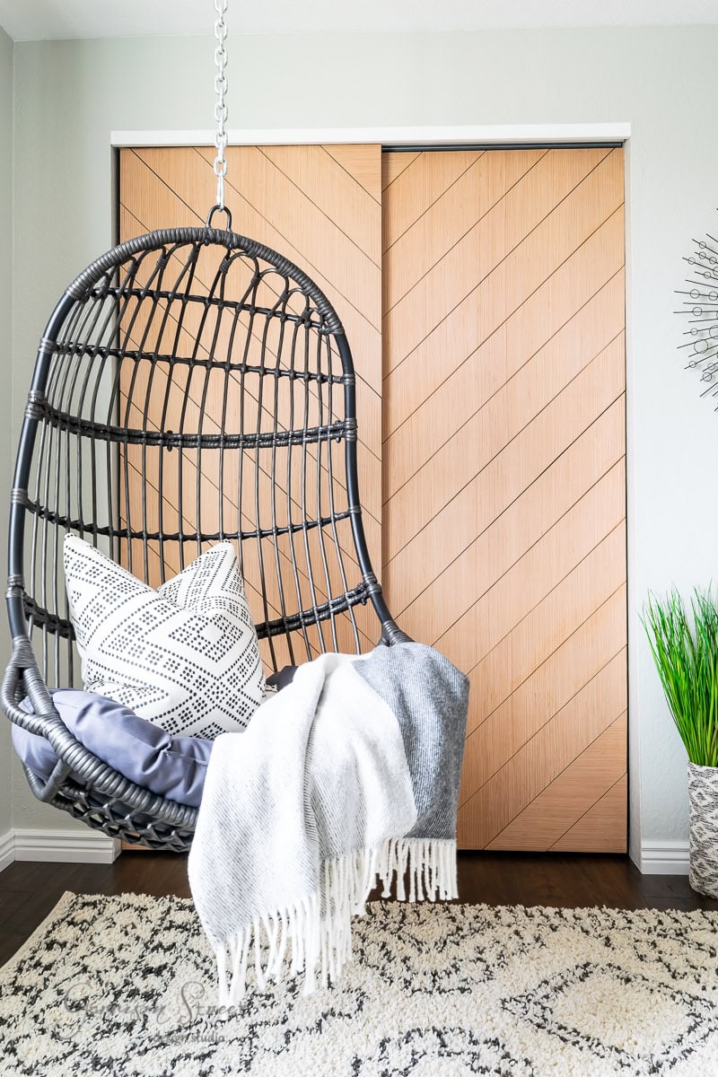 A modern interior space featuring a black hanging wicker chair with a patterned pillow and a gray fringed blanket. In this harmonious project, there's a wooden sliding door with a chevron pattern, a patterned rug, and a potted plant on the floor.