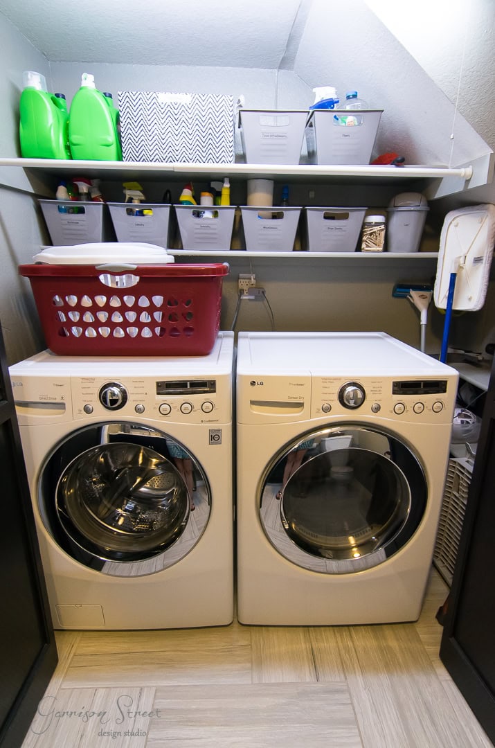 The 2022 laundry room project features a front-loading washing machine and dryer, with shelves above holding bins of cleaning supplies and a red laundry basket. The walls are light gray, complemented by light-colored tile flooring.