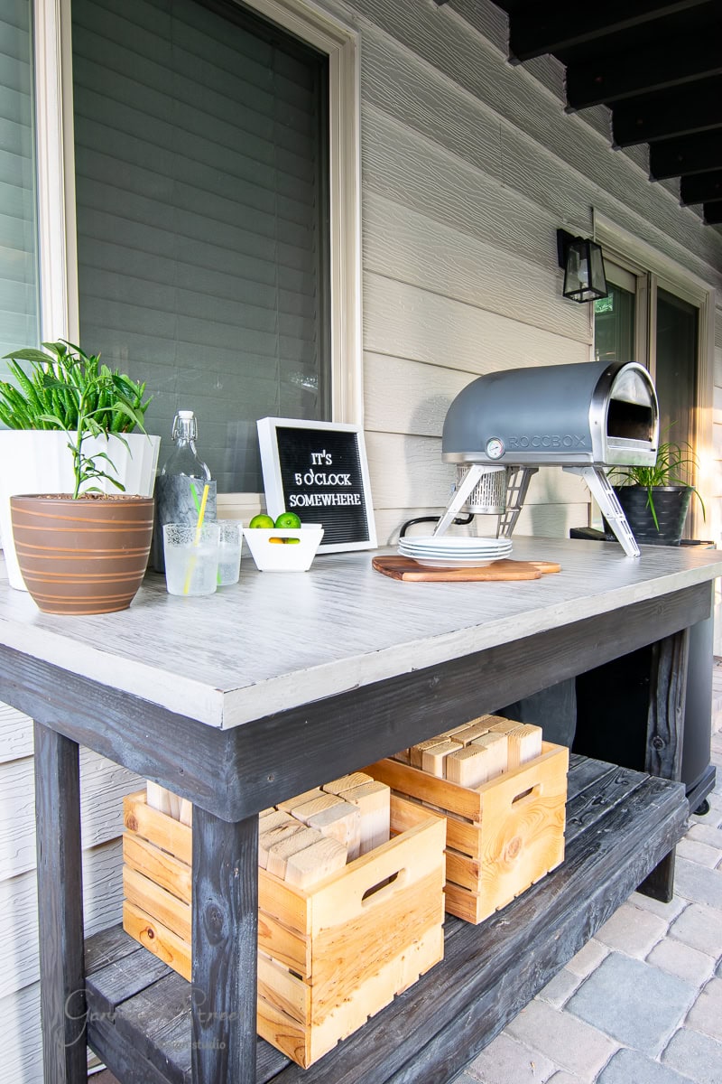 The 2022 patio project features a cozy wooden table setup with a small pizza oven, lush plants, and a refreshing drink with lime. A letter board cheekily declares, "it's 5 o'clock somewhere," while two wooden crates add rustic charm beside the house's wood siding.