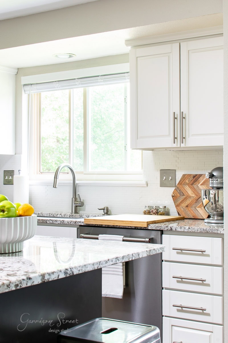 Bright kitchen with white cabinets, stainless steel appliances, and a granite countertop showcasing the perfect setting for culinary projects. A window above the sink lets in natural light, illuminating a bowl of green apples and a wooden cutting board on the counter.