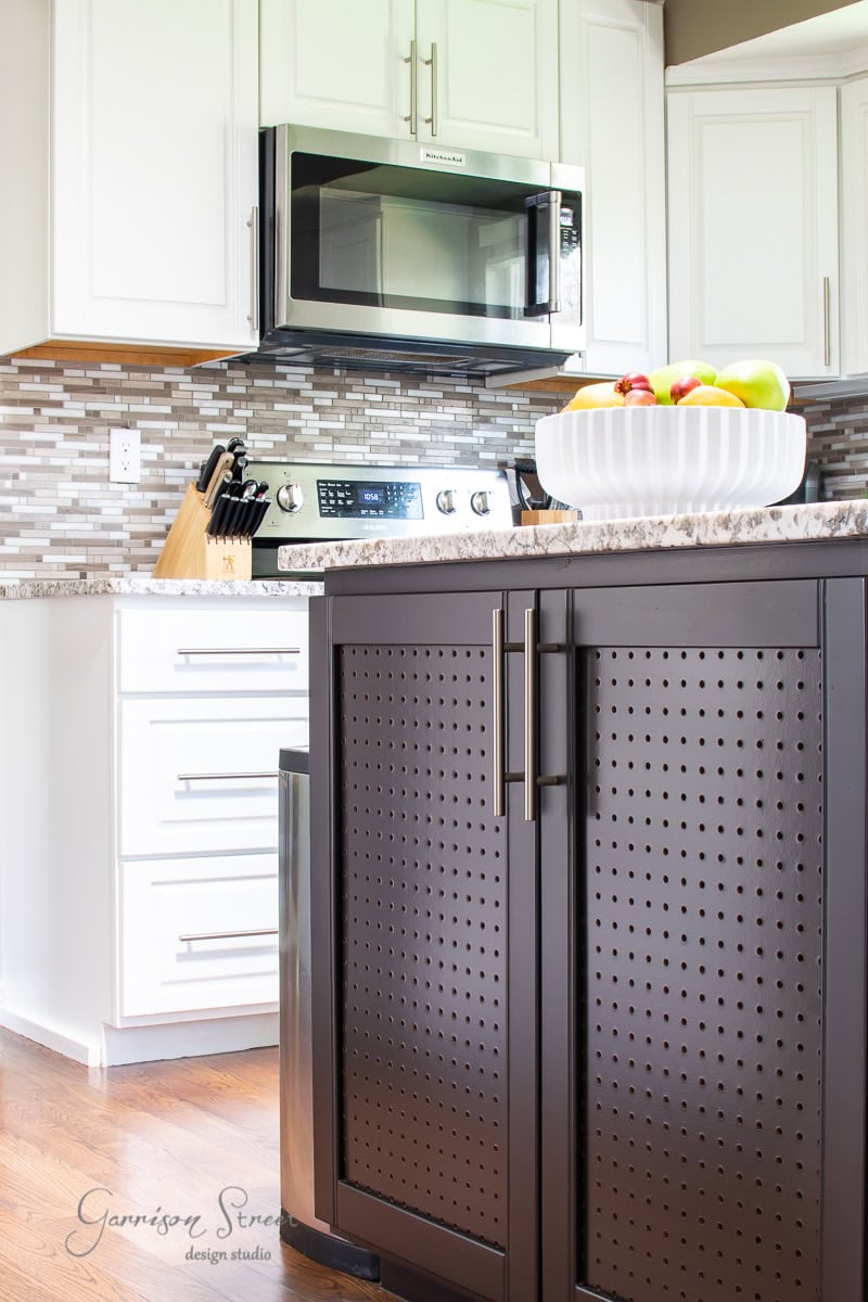 In this modern kitchen, a harmonious blend of white and dark cabinetry frames a stove and microwave. The speckled countertop holds a fruit bowl with apples and bananas, reflecting 2022 design goals. A mosaic tile backsplash and wooden floors complete the project, enhanced by a stylish knife block.