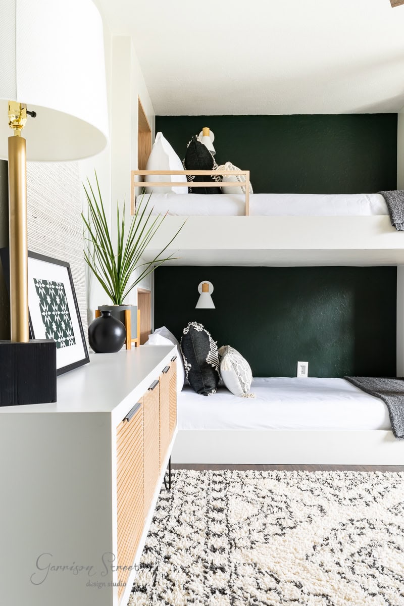 A modern bedroom with white bunk beds against a dark green wall offers a serene setting for your 2022 goals. The top bunk has a small wooden railing and pillows. A patterned floor rug and side table with a lamp and plant add style, enhancing the room's minimalist decor.