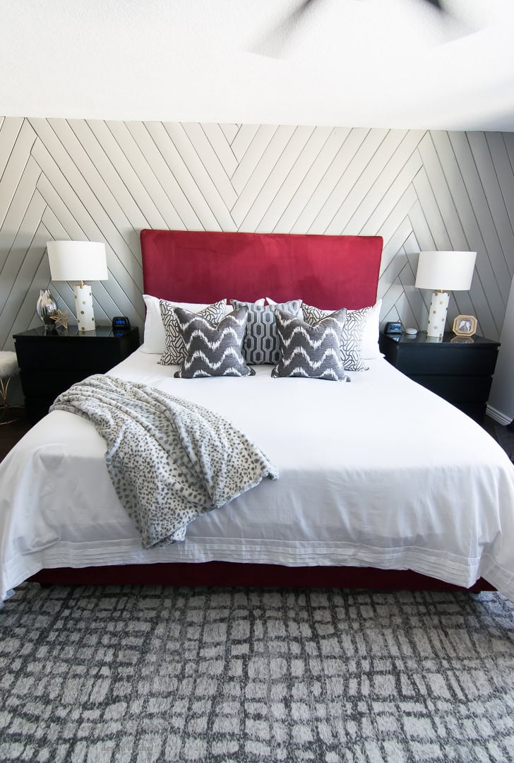 A bedroom with a bed featuring a red headboard, white bedding, and patterned pillows embodies the design goals of 2022. Two bedside tables with lamps and decorative items flank the bed. The wall showcases a chevron pattern, and a textured rug covers the floor.