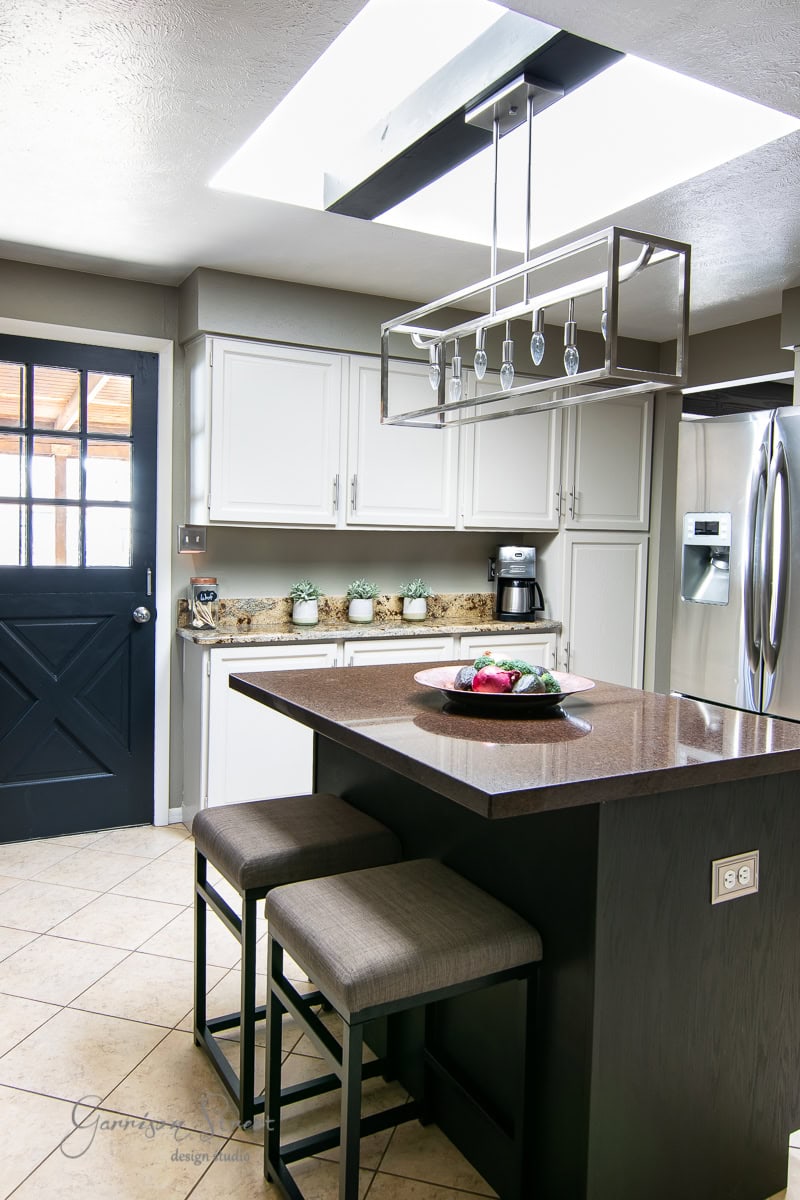 Modern kitchen with white cabinets and stainless steel appliances. In this 2022 project, a dark island with two stools stands at the center, illuminated by a stylish hanging light fixture above. A plate with assorted food sits on the countertop as light streams through the windowed door.