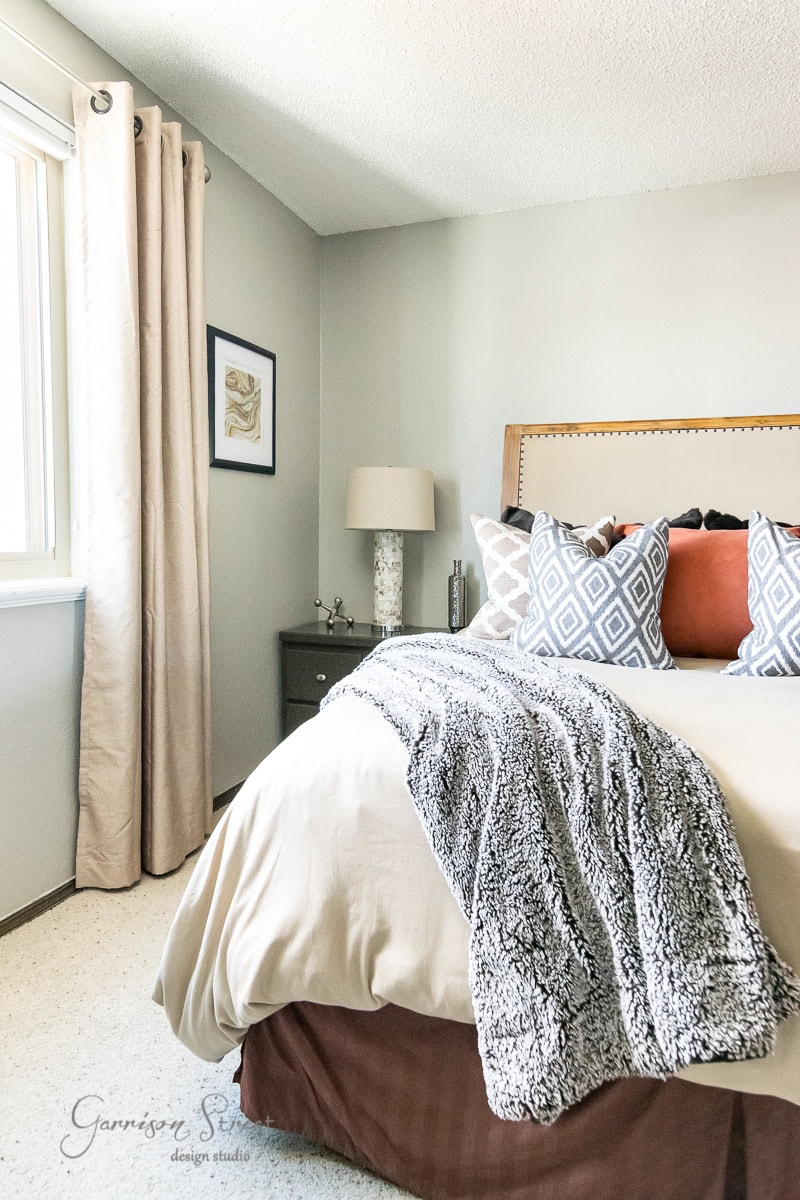 A cozy bedroom with a beige color scheme feels like the perfect retreat to ponder your 2022 goals. The bed is adorned with patterned and solid pillows, along with a fuzzy gray throw blanket. A lamp and framed artwork decorate the nightstand, while light pours in through large windows with long curtains.