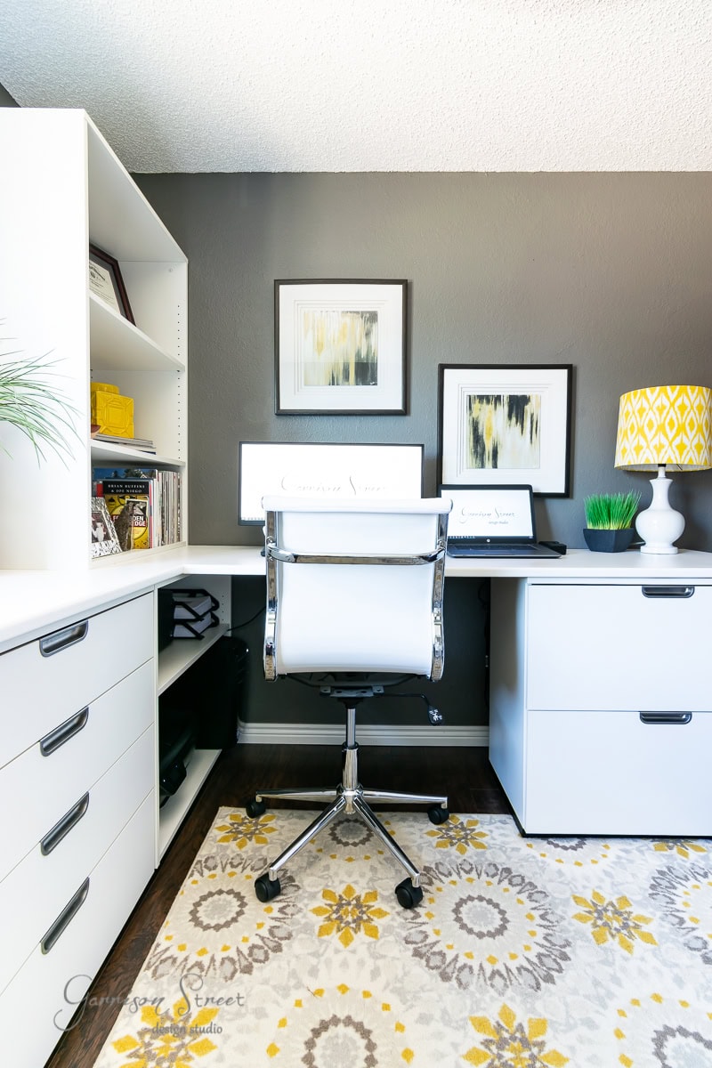A modern home office with a white desk and chair, perfect for tackling 2022 projects. Shelves display books and decor, while two framed abstract artworks adorn the gray wall. A yellow patterned lamp and a plant add color, complementing the yellow and gray floral motif rug beautifully.