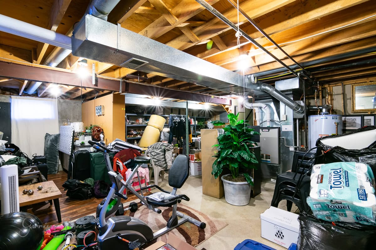 A cluttered basement from 2022, with exposed wooden beams and ductwork, is filled for a project with items like a desk chair, a stationary bike, storage boxes, and a potted plant. Bags of paper towels and miscellaneous household items are scattered around as part of the year’s organization goals.
