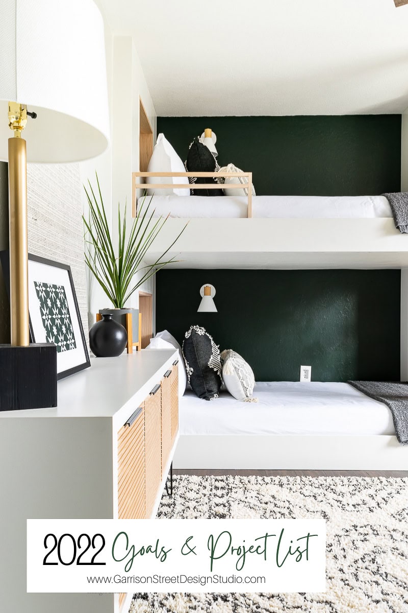 A modern room features white bunk beds against a dark green wall, adorned with simple decorations. Below, a white cabinet holds baskets, decor, framed art, and a plant. A rug covers the floor. Text reads "2022 Project Goals & List", subtly blending style and ambition.