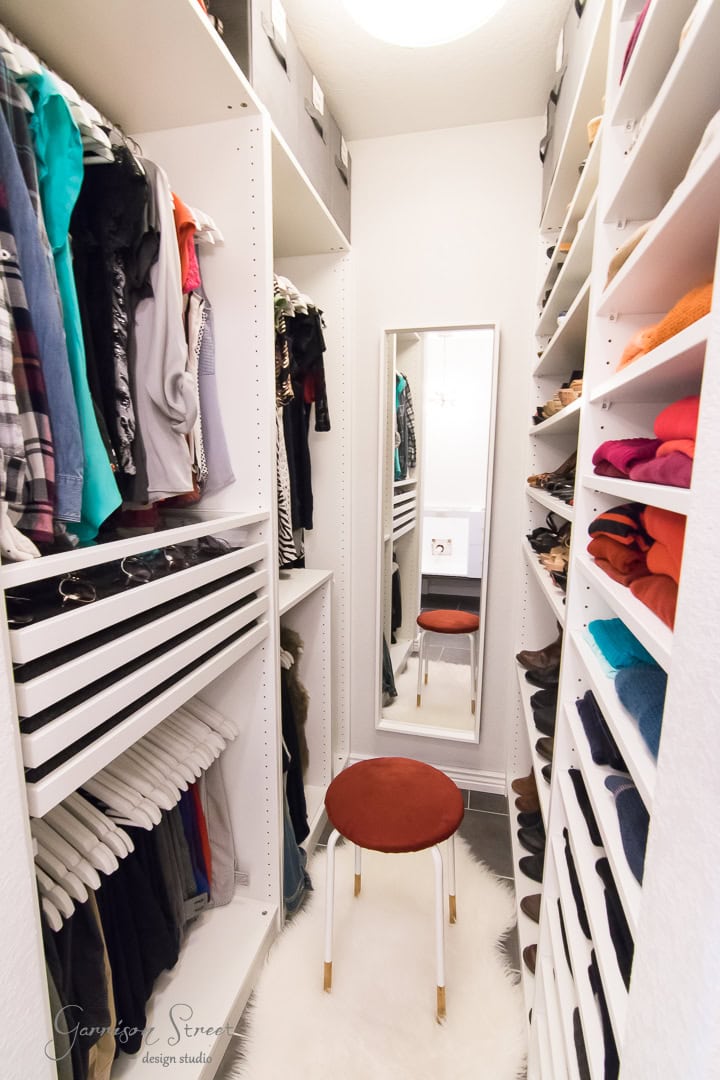 A narrow, organized walk-in closet boasts clothes hanging on both sides, with neatly folded items on shelves and an array of shoes. A full-length mirror stands at the end, reflecting a red stool perfectly placed on a small white rug in the center.
