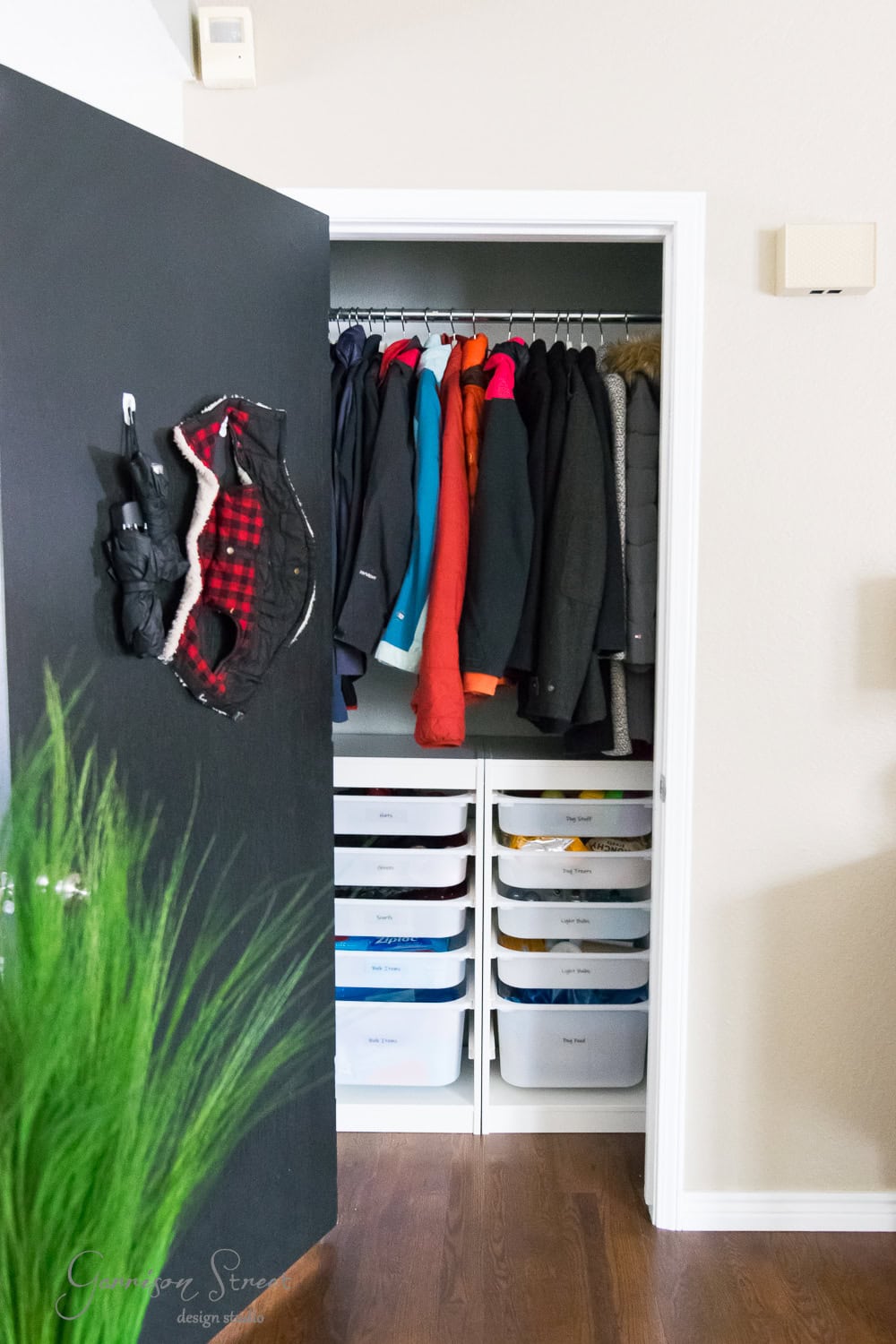 A small closet with a black door is open, showcasing an organized array of hanging coats and two stacks of labeled plastic drawers below. A plant is partly visible in the foreground.