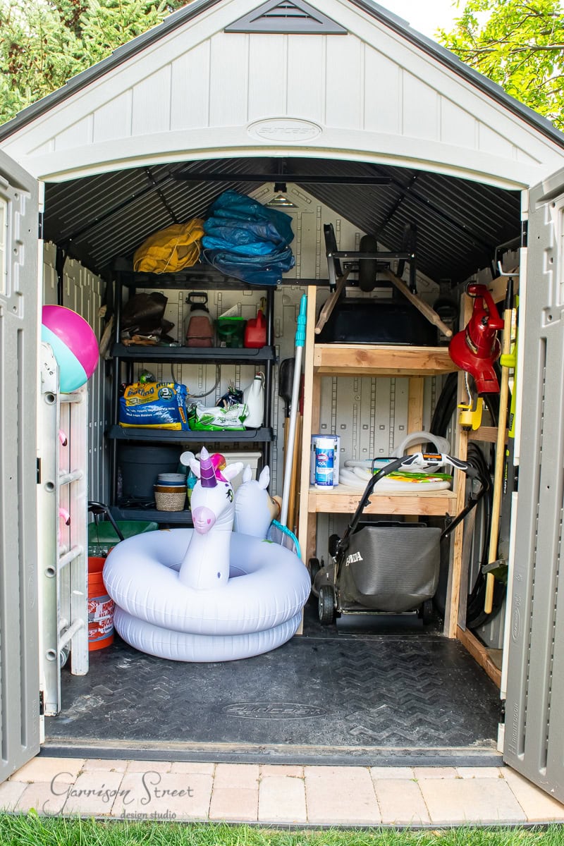 A small shed brimming with garden tools, a lawn mower, neatly organized shelves with paint cans, and a colorful inflatable unicorn pool float. The open shed displays a sense of organization as it reveals its tidy interior.
