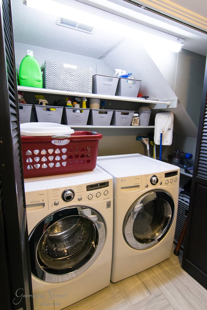 A laundry room with a white washer and dryer side by side exemplifies superb organization. Above them are shelves neatly holding cleaning supplies, storage bins, and laundry detergent. A red laundry basket perches on top of the washer, completing the well-lit and efficiently arranged space.