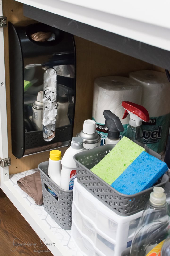 A neatly organized under-sink cabinet brimming with cleaning supplies showcases impeccable organization. Baskets hold sponges, spray bottles, and various cleaning products. On the left side, paper towels and a grocery bag dispenser add convenience in a soothing white and gray color theme throughout.