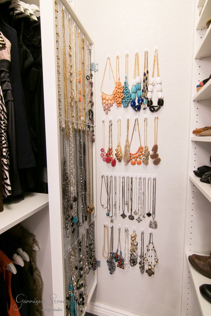 In a closet corner, a white jewelry organizer adds an element of organization to the wall, showcasing necklaces sorted by color. Gold, turquoise, coral, and black pieces catch the eye while adjacent clothing racks complete this stylish setup.