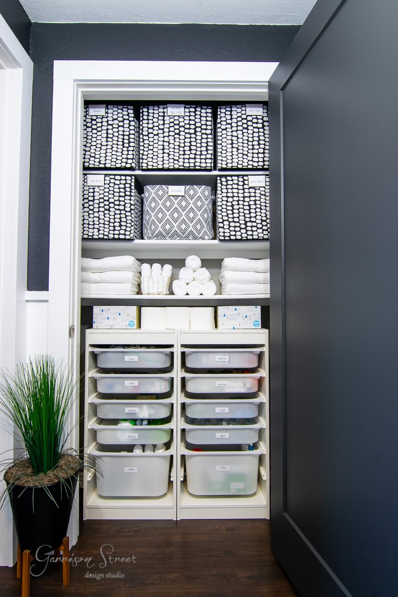 A neatly organized closet showcases shelves with folded towels, patterned baskets, and clear storage bins. Emphasizing organization, a potted plant adds greenery to the space. The dark closet door contrasts beautifully with the white frame.