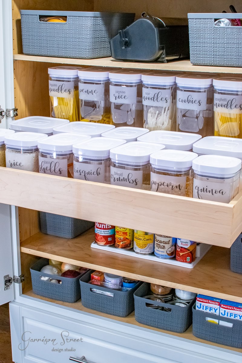 A well-organized pantry shelf showcases labeled clear containers filled with spaghetti, rice, noodles, brown sugar, biscotto, breadcrumbs, popcorn, and quinoa. Exemplifying perfect organization, gray baskets below neatly house canned goods and assorted food packages.
