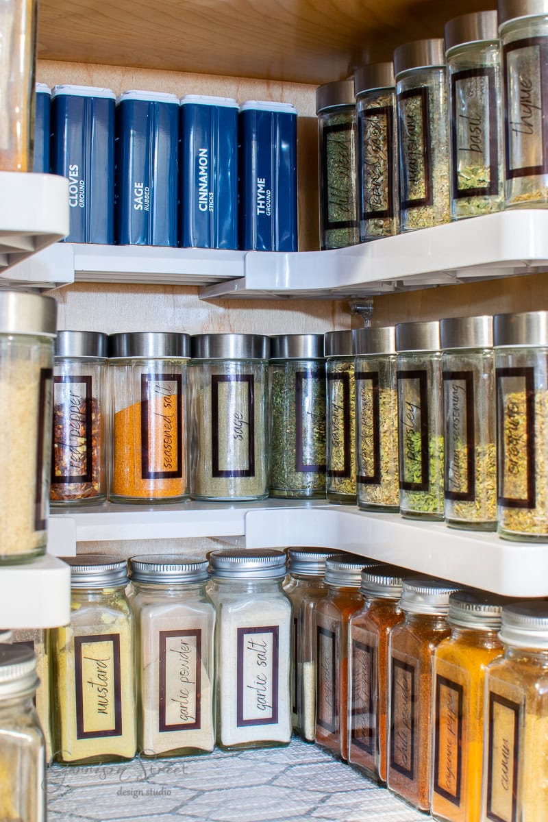 A pantry shelf exhibits impeccable organization, showcasing neatly arranged spices in square glass jars with metal lids. Each jar is labeled, displaying a diverse palette of spices. Larger blue containers hold additional spices on the top shelf.
