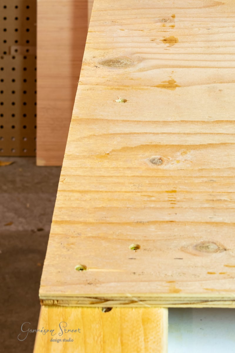 Close-up of a wooden tabletop with visible screws and natural grain patterns, reminiscent of a well-crafted bed frame. The surface is slightly angled, highlighting a neatly finished corner. A perforated board and other wooden pieces are visible in the blurred background.
