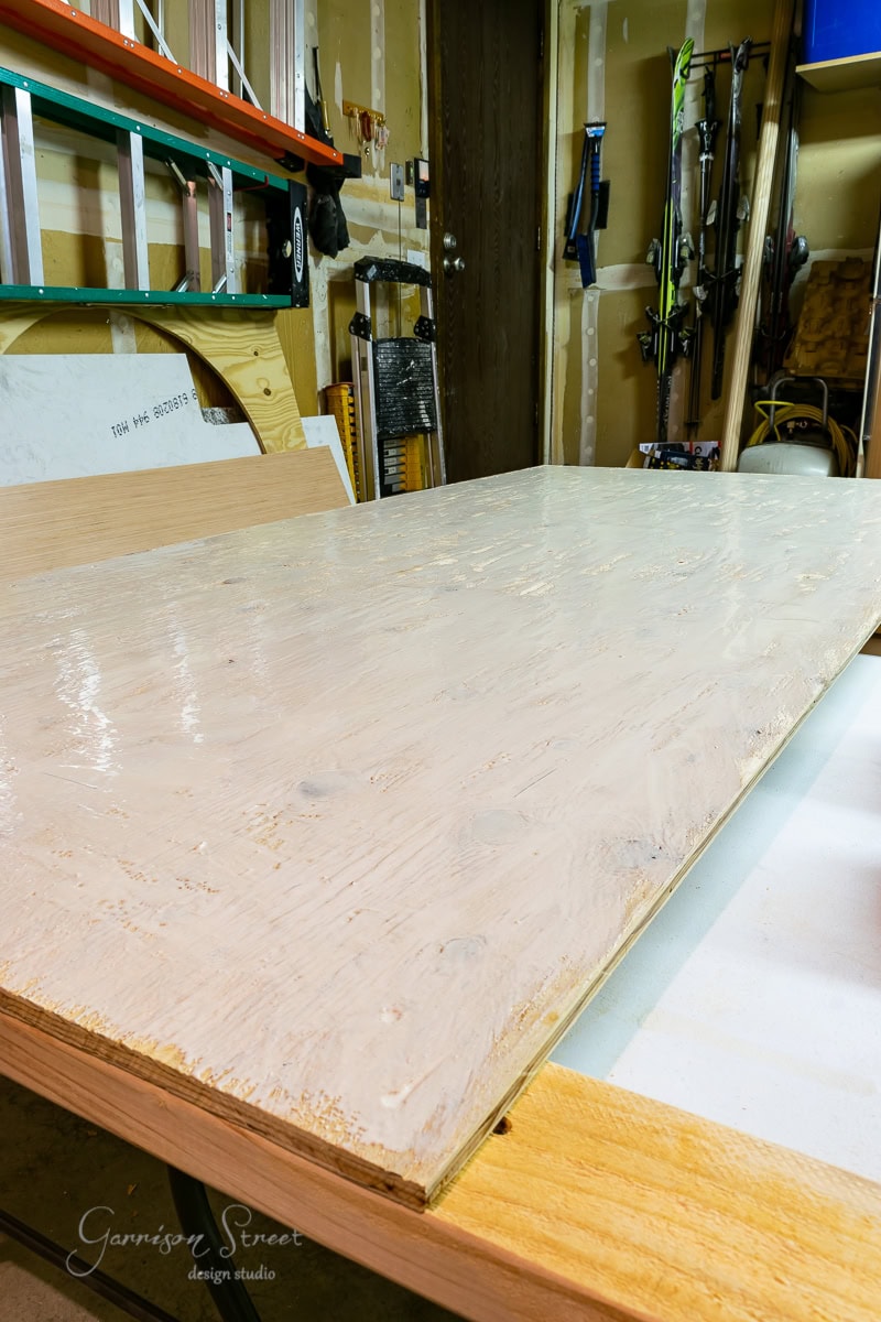 A wooden tabletop in a workshop setting, placed on a workbench with tools and materials visible in the background, including a ladder and various hardware items. Beside it, an adjustable base for a bed frame is ready for assembly. The surface has a light finish.