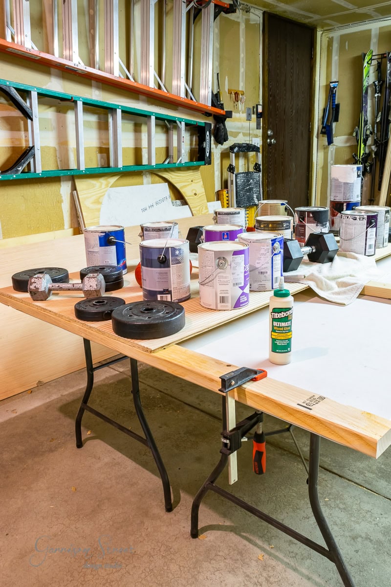 A cluttered garage workbench with several paint cans, weights, and a bottle of Titebond wood glue on a wooden work surface. Ladders and various tools are visible in the background, along with clamps securing a wooden panel that might soon become part of a sturdy bed frame.