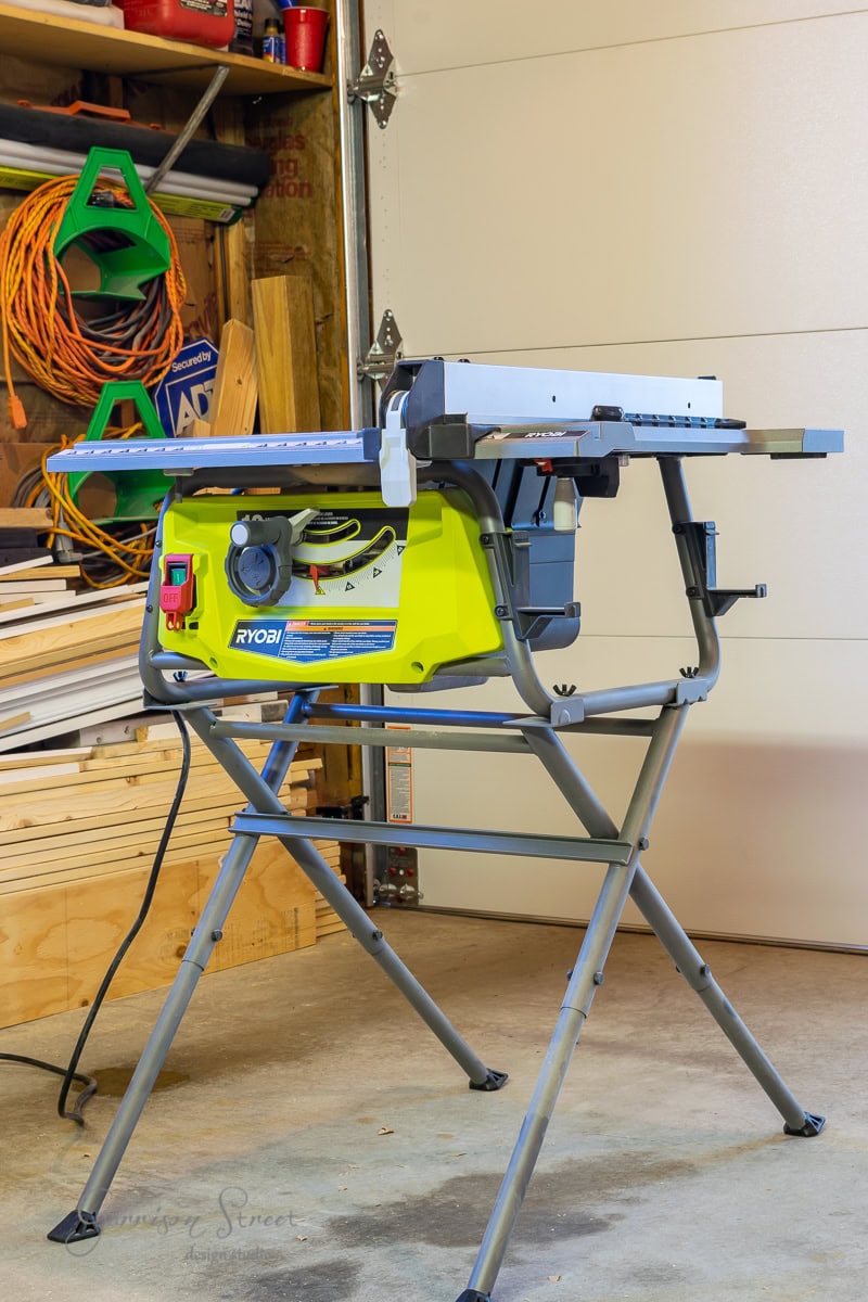 A bright green Ryobi table saw on a metal stand is positioned in a garage, where wooden boards and tools await its craft. Nearby, hangers hold plans for a custom bed frame.