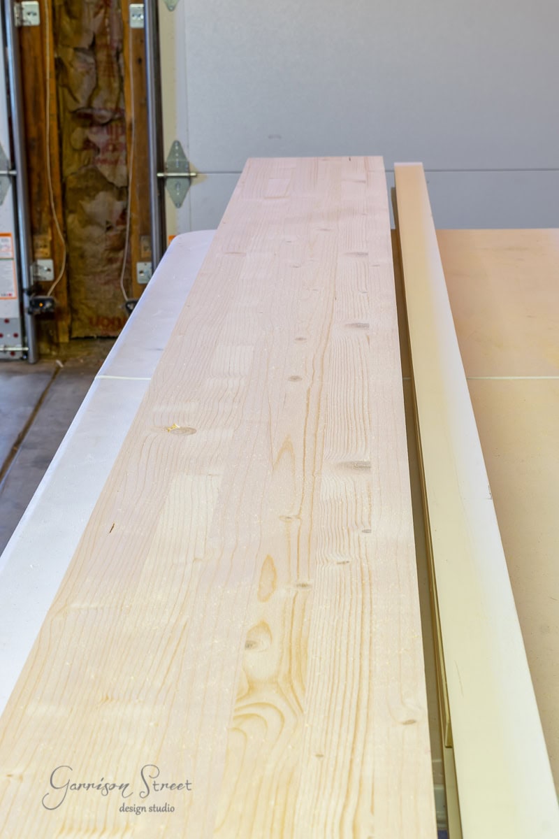 A long wooden board, soon to become part of a stylish bed frame, lies on a flat surface in a workshop. Construction materials are scattered around, hinting at the creativity in progress. A logo in the corner reads "Garrison Street Design Studio.