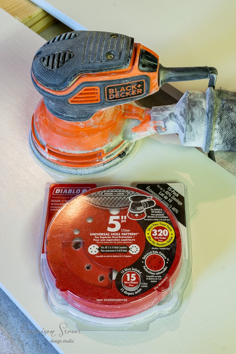 A person using a Black & Decker orbital sander on a bed frame, with a package of Diablo 5-inch sandpaper discs nearby. The sandpaper package highlights features like a universal hole pattern and 220 grit for polishing, perfect for achieving that flawless finish on your adjustable base.