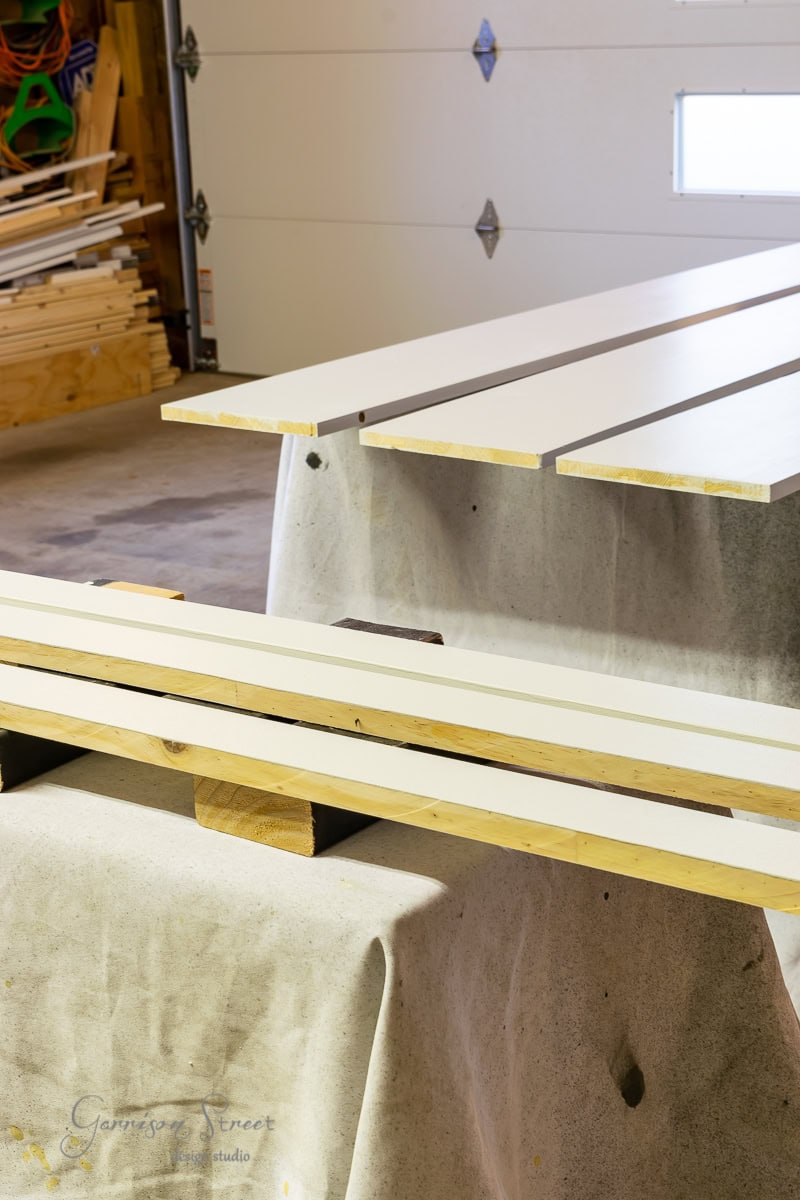 Long wooden planks, soon to become part of a custom bed frame, dry on a table covered with cloth in a workshop. The background is filled with more lumber and a partially open garage door, hinting at an adjustable base under construction.