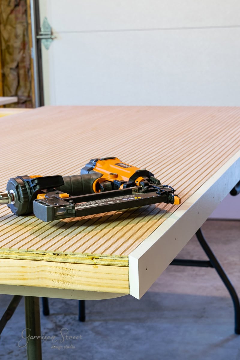 A nail gun sits atop a striped, unfinished wooden board, perhaps destined to form part of a sturdy bed frame. The setting resembles a workshop or garage, with an adjustable base and other tools visible in the background, hinting at ongoing projects.