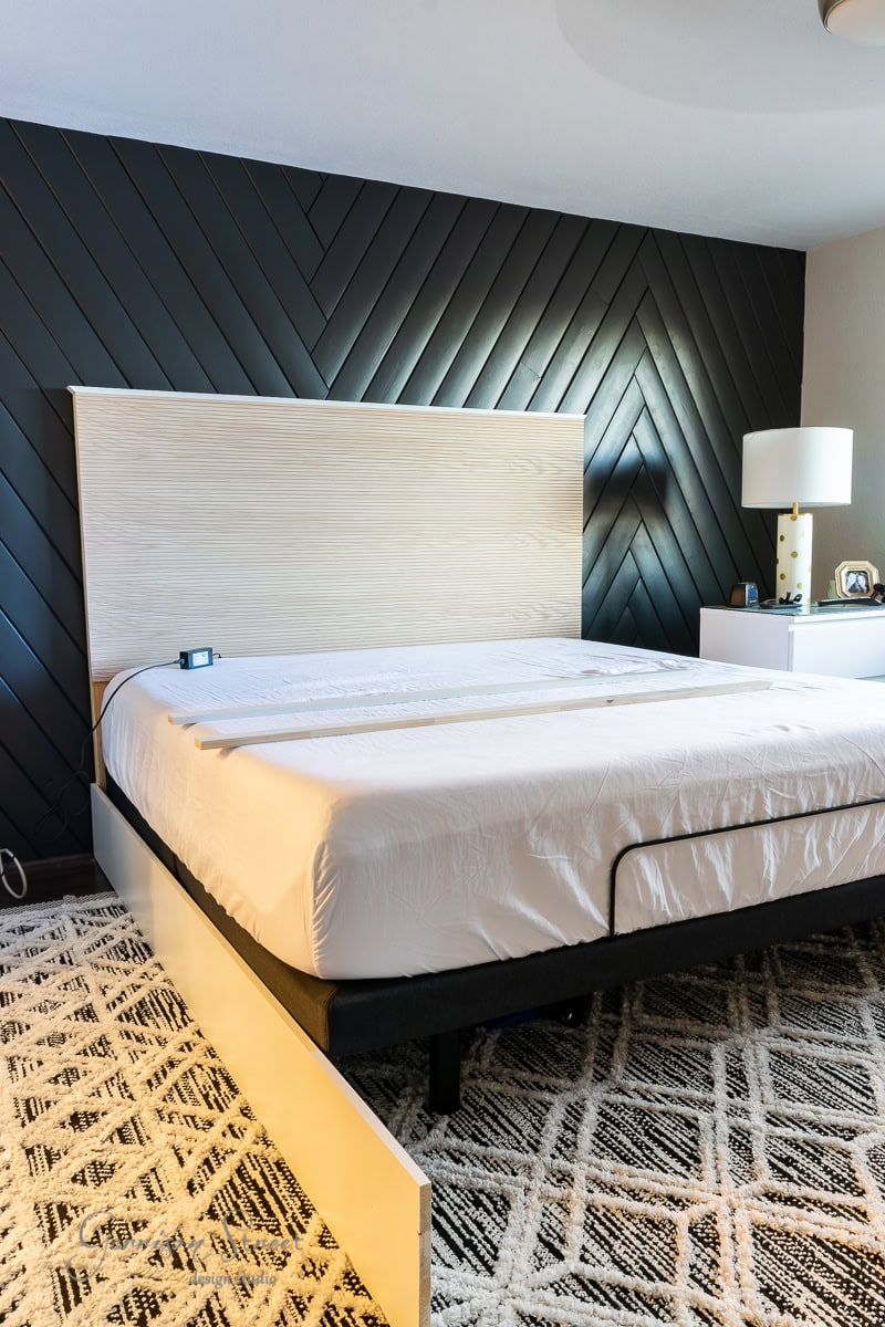 A modern bedroom showcases a bed with a light wood adjustable base headboard against a dark herringbone accent wall. A white nightstand with a lamp and small clock sits beside the bed frame, and the floor features a patterned rug.