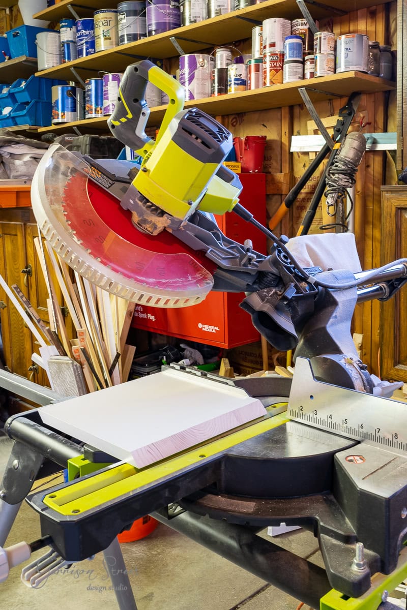 A green miter saw sits on a workbench in a cluttered workshop, poised over a piece of white wood destined for an adjustable base project. Shelves filled with paint cans and tools create a colorful backdrop, adding character to the creative chaos.